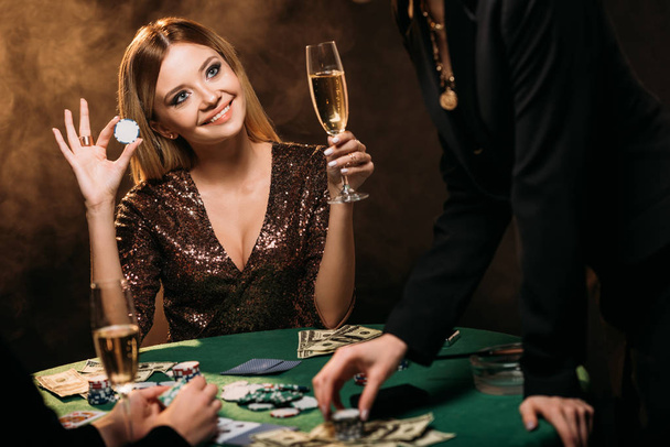 beautiful smiling woman holding glass of champagne and poker chip at table in casino - Photo, Image