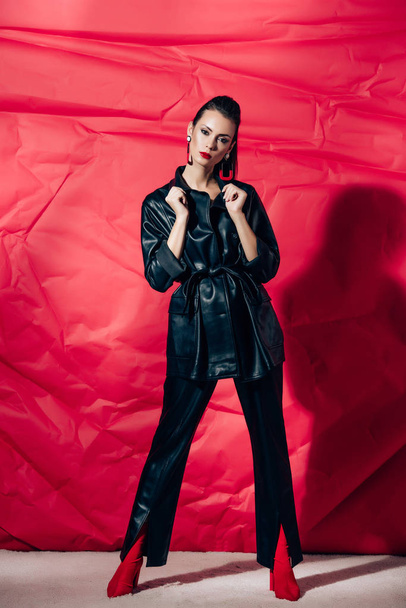 attractive fashionable young woman posing in black leather suit on red background - Foto, Imagem