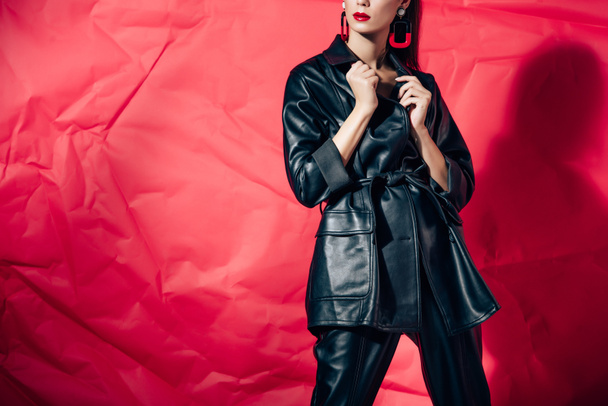 cropped view of stylish woman posing in black leather suit on red background - 写真・画像