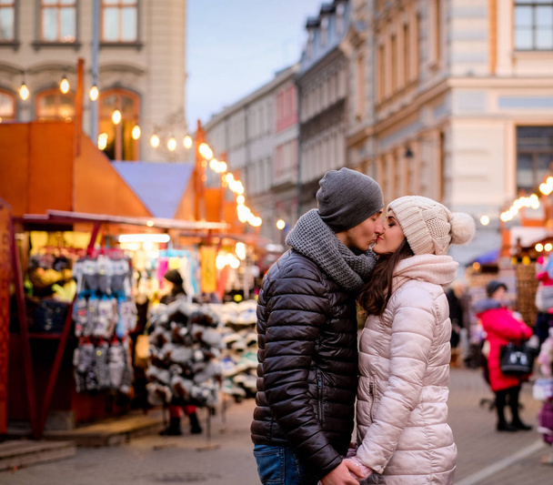 Mladý romantický pár na sobě zimní oblečení, chyťte se za ruce a líbání při stání v ulici večer s vánoční jarmark - Fotografie, Obrázek