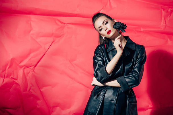 beautiful model in black leather suit posing with black rose flower on red background - Fotó, kép