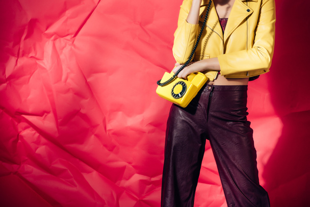 cropped view of woman in yellow leather jacket posing with rotary phone on red background - Photo, Image