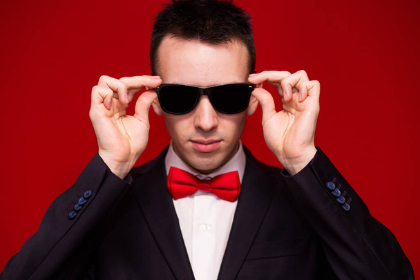 Porträt eines ernst zu nehmenden gutaussehenden, stilvollen Mannes in elegantem schwarzen Anzug und Sonnenbrille auf rotem Hintergrund. Geschäftsstil. Modisches Image - Foto, Bild
