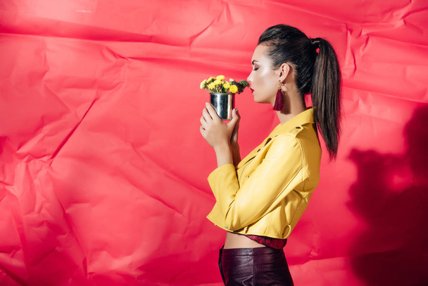 side view of fashionable girl in yellow leather jacket posing with flowers on red background - Фото, изображение
