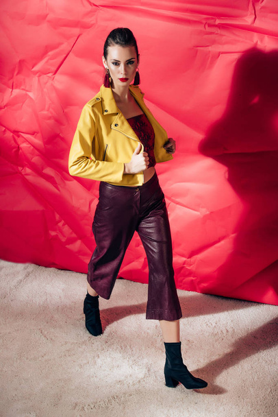 stylish model posing in yellow leather jacket on red background for fashion shoot - Foto, Imagem