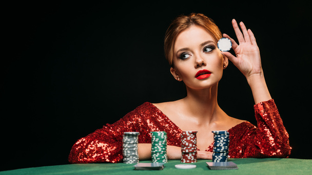 attractive girl in red shiny dress leaning on table, holding poker chip and looking away isolated on black - Fotoğraf, Görsel