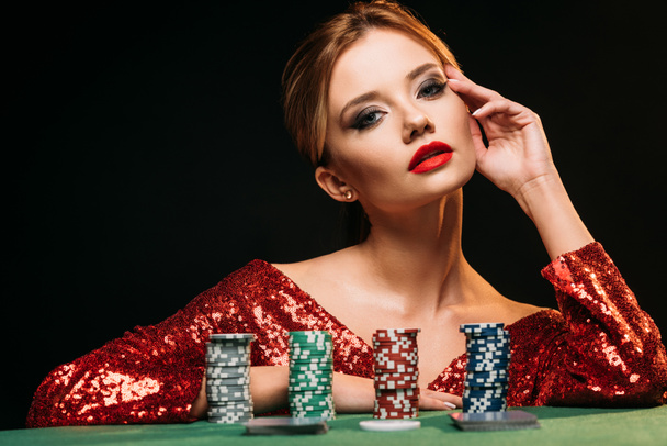 beautiful girl in red shiny dress leaning on table with poker chips and looking at camera isolated on black - Photo, Image
