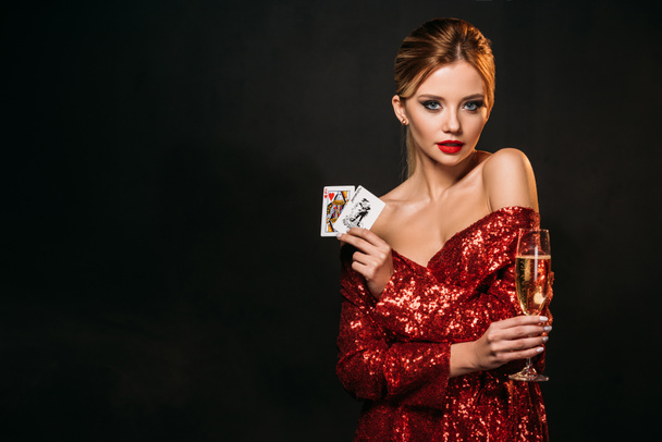 attractive girl in red shiny dress holding joker and queen of hearts cards isolated on black, looking at camera - Photo, image