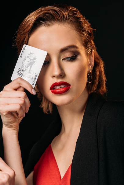 portrait of attractive girl in red dress and black jacket holding joker card isolated on black - Photo, Image