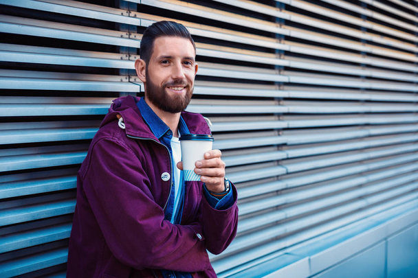 Handsome young man in purple winter jacket drinking coffee. The guy drinking coffee on the street. - Photo, Image