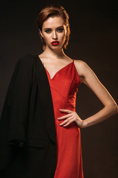 красива жінка в червоній сукні і чорна куртка на одному плечі дивиться на камеру ізольовано на чорному
 - Фото, зображення