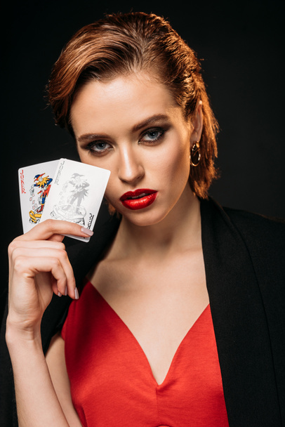 portrait of attractive girl in red dress and black jacket holding poker cards and looking at camera isolated on black - Photo, Image
