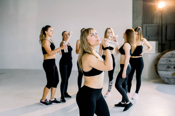 Group of young sporty girls with water boottles standing at white wall. Female companions in gym resting after fitness, indoor full length, loft studio, copy space - Foto, imagen