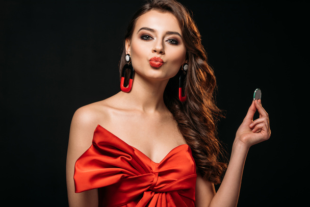 beautiful brown haired girl in red corset holding casino chip and pretending kissing isolated on black - Фото, изображение
