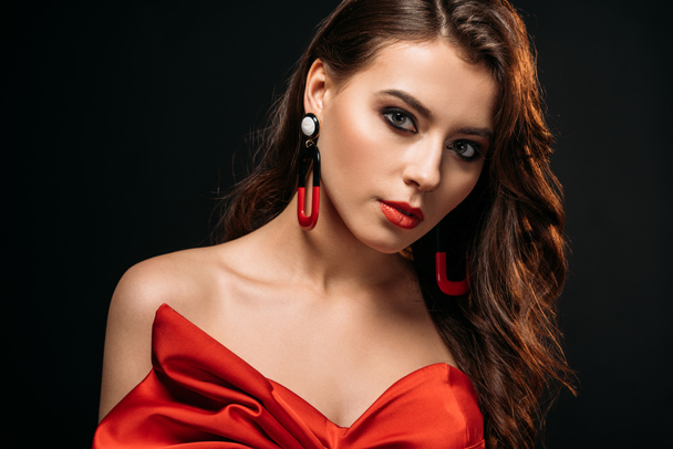 portrait of beautiful brown haired girl in red corset and earrings looking at camera isolated on black - Foto, immagini