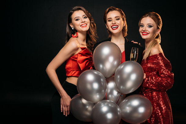 smiling attractive girls in stylish party clothes holding bundle of grey balloons and looking at camera isolated on black - Foto, Imagem