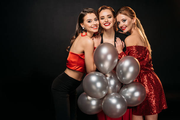 smiling attractive girls in stylish party clothes holding bundle of grey balloons, hugging and looking at camera isolated on black - Photo, Image
