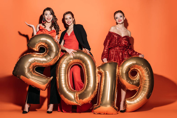 surprised attractive girls in stylish party clothes holding 2019 balloons and looking at camera on orange - Foto, afbeelding