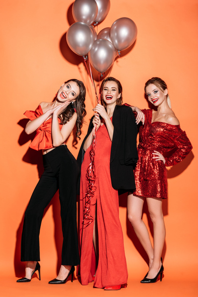 excited beautiful women in stylish red and black clothes holding bundle of grey balloons and looking at camera on orange - Foto, Bild
