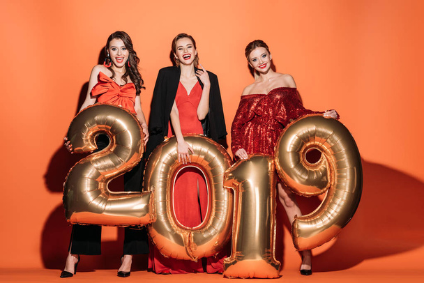 Улыбающиеся привлекательные девушки в стильной одежде для вечеринок с воздушными шарами 2019 года на апельсине
 - Фото, изображение