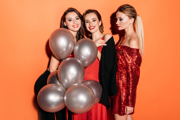 smiling attractive girls in stylish party clothes holding bundle of grey balloons isolated on orange - Foto, Imagen