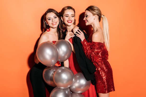 smiling attractive girls in stylish party clothes holding bundle of grey balloons isolated on orange - Foto, immagini