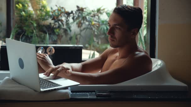 Νεαρός άνδρας βρίσκεται στο μπάνιο και λειτουργεί με έναν φορητό υπολογιστή - Πλάνα, βίντεο