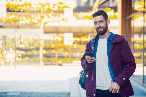 Hombre barbudo confiado sosteniendo y teléfono móvil mientras está de pie cerca de la pared de la calle en la soleada noche de verano
. - Foto, imagen