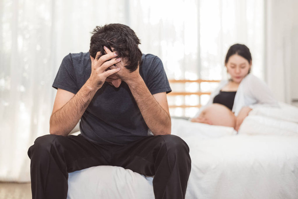 Worried stress man sitting on bed with hand on forehead in bedroom in serious mood emotion with pregnant wife woman background. Major Depressive Disorder called MDD concept. Physical healthcare - Photo, Image