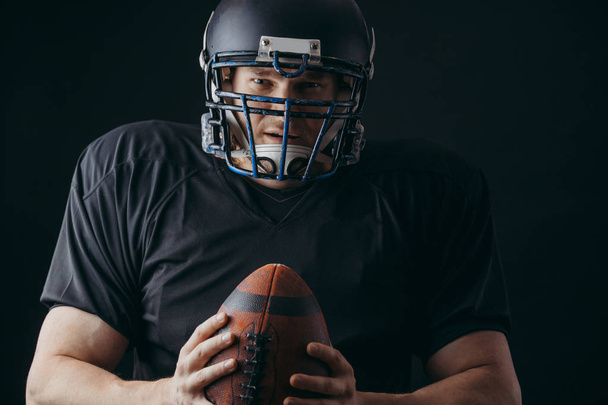 American football player in black sportwear with a ball on black background - Фото, зображення