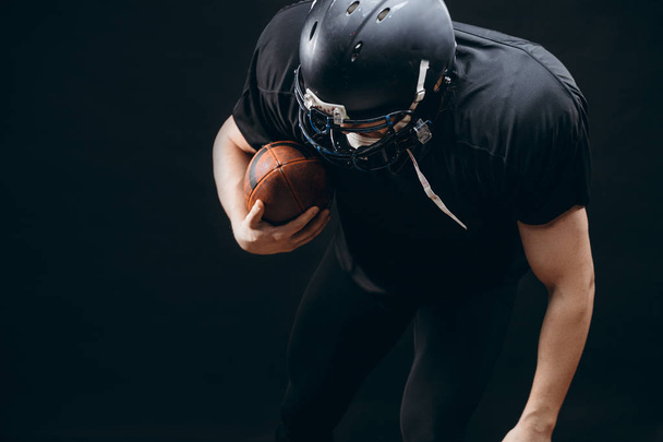American football player with helmet and armour with a ball against black wall - Foto, immagini