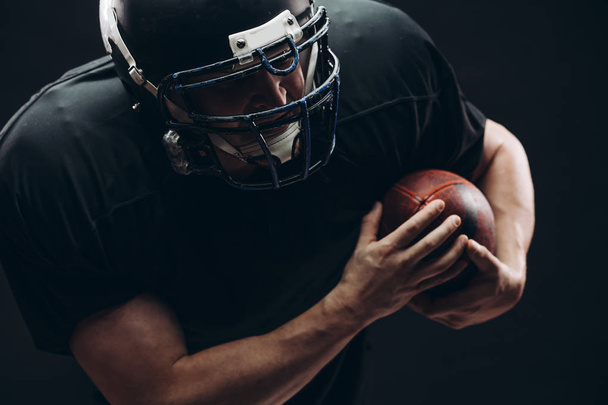American football player with helmet and armour with a ball against black wall - Foto, Bild