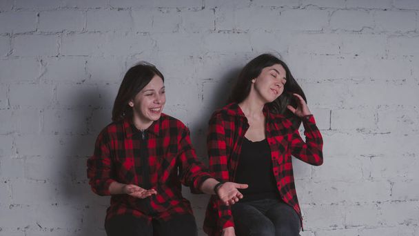 Two young girls in red and black plaid shirts sitting on chairs on brick wall background, female friendship concept - Photo, image