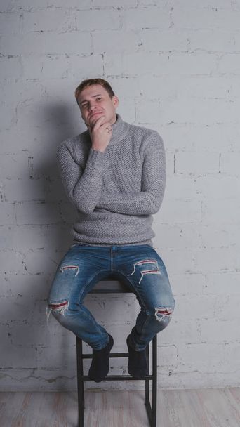 The guy in the warm sweater on the chair, on a gray background. A man posing against a brick wall. - Foto, imagen