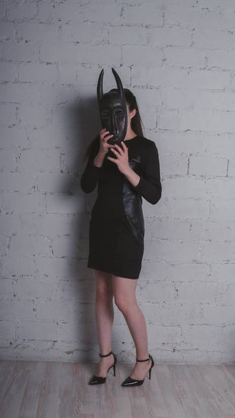 Photo of a young brunette in a black dress, with an ancient mask, against a white brick wall in the Studio - Photo, image