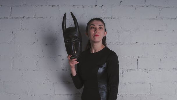 Photo of a young brunette in a black dress, with an ancient mask, against a white brick wall in the Studio - Foto, Imagem