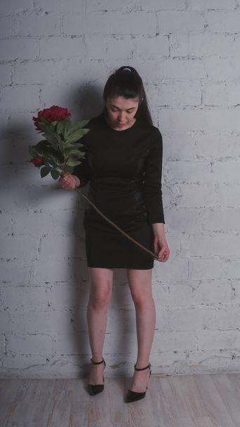A slender girl is standing in a black dress, holding a large artificial flower and posing against a gray wall - Photo, Image