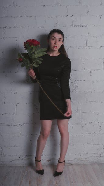A slender girl is standing in a black dress, holding a large artificial flower and posing against a gray wall - Photo, image