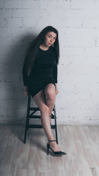 Girl posing against a white brick wall. Model in black dress. Brunette Eastern looks. - Фото, изображение