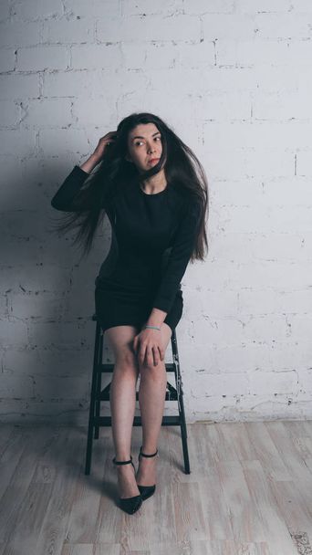 Girl posing against a white brick wall. Model in black dress. Brunette Eastern looks. - Фото, зображення