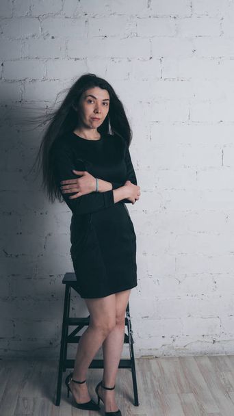 Girl posing against a white brick wall. Model in black dress. Brunette Eastern looks. - Fotó, kép