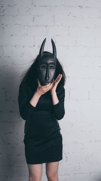 Photo of a young brunette in a black dress, with an ancient mask, against a white brick wall in the Studio - Fotografie, Obrázek