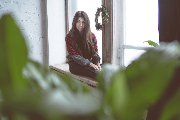 Mädchen am Fenster sitzend, eine Studentin im grauen Pullover. Modell gegen das Fenster posiert. Konzept: Romantik und Sehnsucht - Foto, Bild
