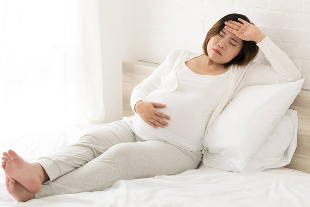 Asiática embarazada con la mano en la frente sufriendo dolor de cabeza sentado en la cama en casa
 - Foto, Imagen