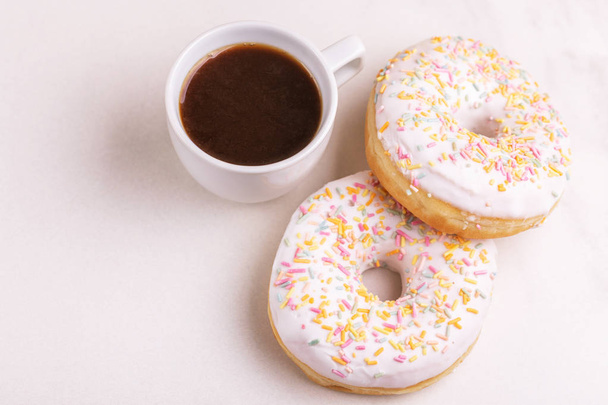 Two delicious vanilla donuts with sprinkle and cup of coffee on light marble background. Unhealthy, but tasty sweets. Copy space. Horizontal view - Foto, Bild