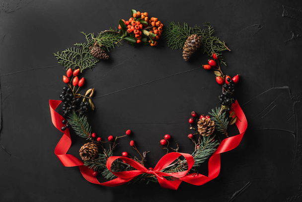 Crown of Christmas tree branches, pine cones, berries, and red slack, on black stone background. Xmas and Happy New Year theme. Flat lay, top view - Foto, Imagem