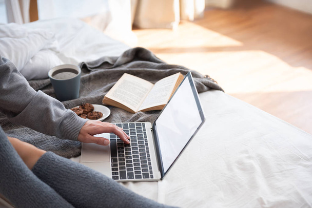 Nő kéz a szürke pulóver segítségével és vele laptop gépelés ülve, ágyban otthon. Pihentető ázsiai nő, egy csésze tea és a cookie-kat a nyaralás. Másolja a hely. - Fotó, kép