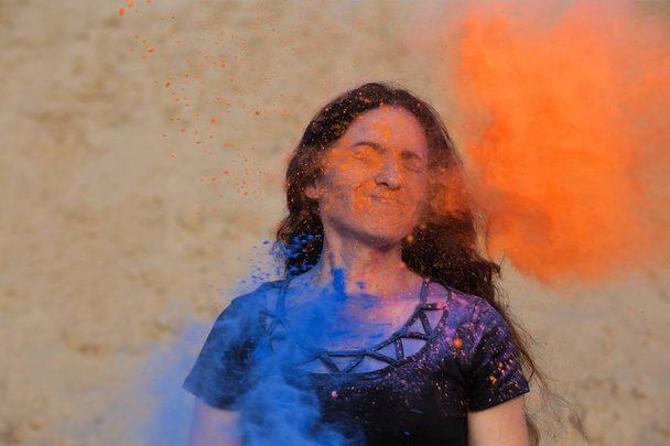 Pretty young model posing with exploding around her blue and orange Holi powder at the desert - Photo, Image