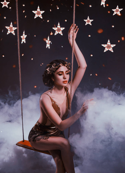 Girl star riding on a swing in the night sky above the clouds. Greek goddess in a golden dress and wreath. Art photo - Foto, Bild