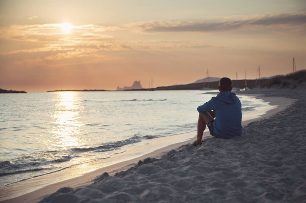 ein junger Mann sitzt am Strand im Sand und blickt in die Ferne. - Foto, Bild